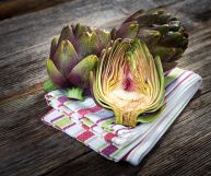 Su uno strofinaccio bianco a righe rosse, In primo piano, carciofo tagliato; dietro due ortaggi interi; sotto un asciugamano da cucina su un tavolo in legno