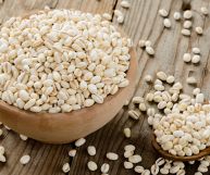 Da sinistra a destra, una ciotola di legno chiaro e un cucchiaio sempre in legno ma scuro, entrambi riempiti con chicchi d'orzo color panna. Tavolo marrone.