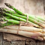Due mazzi di asparagi freschi e puliti, ma con ancora la parte fibrosa; sono sia verdi che bianchi e posati su un rettangolo di iuta su tavolo rustico
