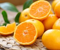 Arance di colore arancione chiaro; per la maggior parte sono intere, alcune sono tagliate a metà; sono posate su tovaglietta in vimini e tavolo di legno