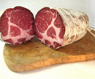 Su un tagliere di legno, due metà di un capocollo calabrese dal tipico colore rosato e rossastro con striature bianche di grasso; lo sfondo è bianco