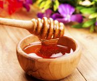 Uno bastoncino per mescolare il miele intriso di miele marrone- dorato che cola in una scodella di legno chiaro; piano in legno e fiori colorati sullo sfondo