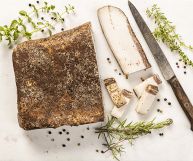 Su uno sfondo bianco, da sx a sx, pezzo di lardo di Colonnata quasi intero, cosparso di spezie, una fetta bianca e un coltello con manico nero e lama tagliente;