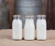 Latticello liquido contenuto in tre bottiglie di vetro tutte uguali e senza tappo, posate in fila su un piano in legno. Lo sfondo è scuro, tendente al grigio