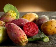 Un frutto tagliato a metà e alcuni fichi d'India di colore rosso brillante, giallo senape e arancione chiaro posati su un piano rustico di legno