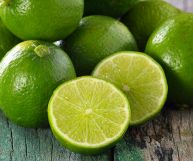 Lime interi e tagliati a metà appoggiati su un tavolo di legno. Hanno forma rotonda, una sottile buccia verde brillante e una polpa più chiara
