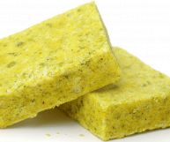 Su sfondo bianco, due tavolette di dado di verdure senza carta; presentano il tipico colore giallognolo-verdastro dei prodotti industriali