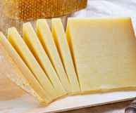 Davanti pezzo e fette più sottili di groviera con crosta chiara; sullo sfondo pezzo con scorza più scura; sulla destra coltello con manico in legno