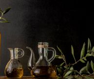 Sulla sinistra tre bottiglie contenenti olio evo dal colore intenso; sono di vetro, con forme e altezze diverse. Sulla destra ramoscello di olivo
