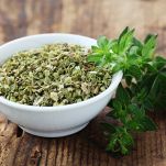 Una ciotola bianca contenente origano essiccato pronto all’uso, affiancata da un ramoscello di origano fresco; il tutto appoggiato su un piano di legno