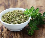 Una ciotola bianca contenente origano essiccato pronto all’uso, affiancata da un ramoscello di origano fresco; il tutto appoggiato su un piano di legno