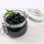 Olive nere dalla polpa soda e lucida, pronte per essere gustate. Sono contenuto in un piccolo barattolo di vetro con coperchio aperto. Sfondo chiaro