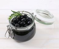 Olive nere dalla polpa soda e lucida, pronte per essere gustate. Sono contenuto in un piccolo barattolo di vetro con coperchio aperto. Sfondo chiaro