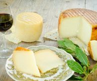 Una forma cilindrica di pecorino sardo da cui sono state prelevate due spesse fette posate su un piatto trasparente; dietro un calice di vino rosso