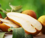 Davanti due metà di pere Abate dalla polpa bianca e croccante; dietro pere intere e mature, dalla buccia giallo-aranciata; tavolo color panna e sfondo verde