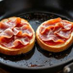 Fette di speck adagiate su metà panino su piastra calda.