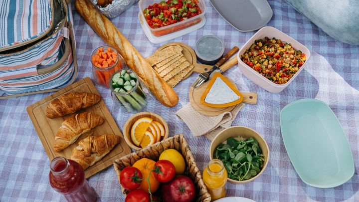 Allestimento per picnic su una tovaglia a quadri con una borsa termica a righe sulla sinistra, baguette, croissant, frutta fresca e insalate in contenitori.
