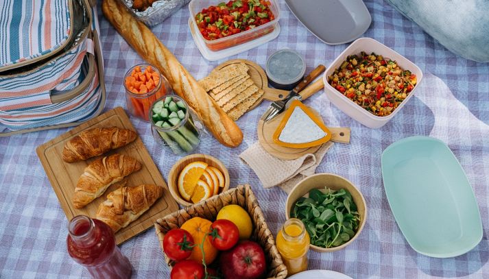 Allestimento per picnic su una tovaglia a quadri con una borsa termica a righe sulla sinistra, baguette, croissant, frutta fresca e insalate in contenitori.