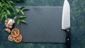 Coltello da chef professionale con manico nero posato su un fondo scuro, accanto a un tagliere in ardesia rettangolare vuoto, aglio e spezie.