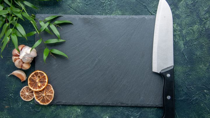 Coltello da chef professionale con manico nero posato su un fondo scuro, accanto a un tagliere in ardesia rettangolare vuoto, aglio e spezie.