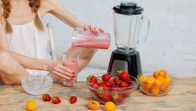 Donna che versa uno smoothie di fragole e albicocche in un bicchiere, con ciotole di frutta fresca e ghiaccio sul tavolo.