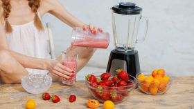 Donna che versa uno smoothie di fragole e albicocche in un bicchiere, con ciotole di frutta fresca e ghiaccio sul tavolo.