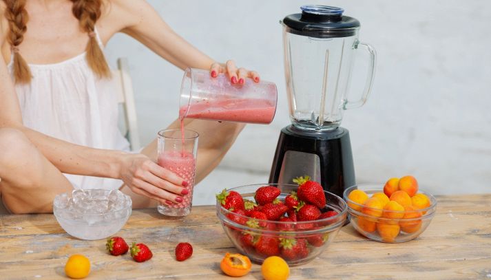 Donna che versa uno smoothie di fragole e albicocche in un bicchiere, con ciotole di frutta fresca e ghiaccio sul tavolo.