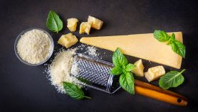 Composizione flat lay con grattugia manuale tipo microplane, scaglie di parmigiano e formaggio grattugiato fresco su sfondo scuro con basilico.