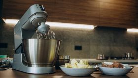 Impastatrice compatta con frusta e ciotola inox su piano cucina, ingredienti pronti per l’impasto.