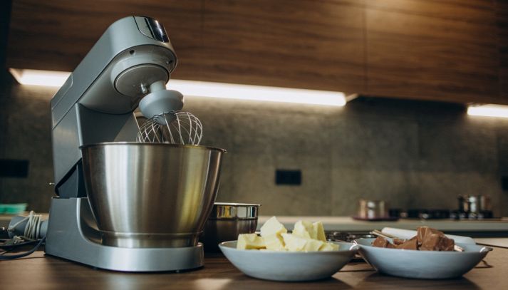 Impastatrice compatta con frusta e ciotola inox su piano cucina, ingredienti pronti per l’impasto.