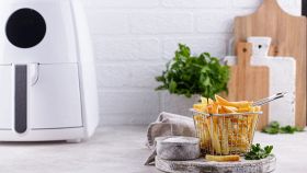 Cestello in metallo colmo di patatine fritte dorate posizionato davanti a una mini air fryer bianca, su un piano cucina con sale e prezzemolo.