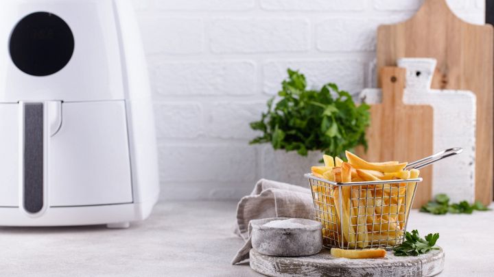 Cestello in metallo colmo di patatine fritte dorate posizionato davanti a una mini air fryer bianca, su un piano cucina con sale e prezzemolo.