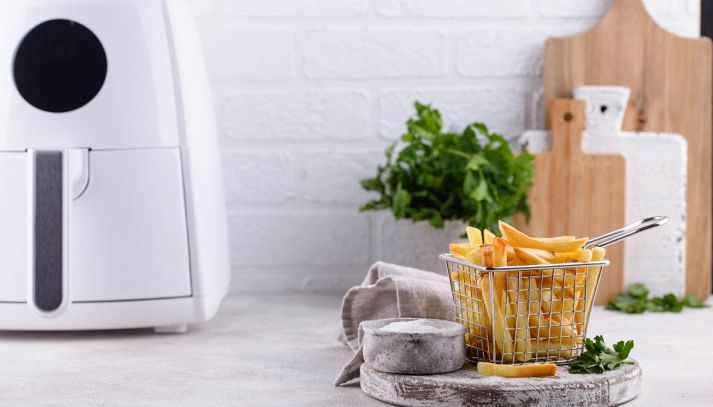 Cestello in metallo colmo di patatine fritte dorate posizionato davanti a una mini air fryer bianca, su un piano cucina con sale e prezzemolo.