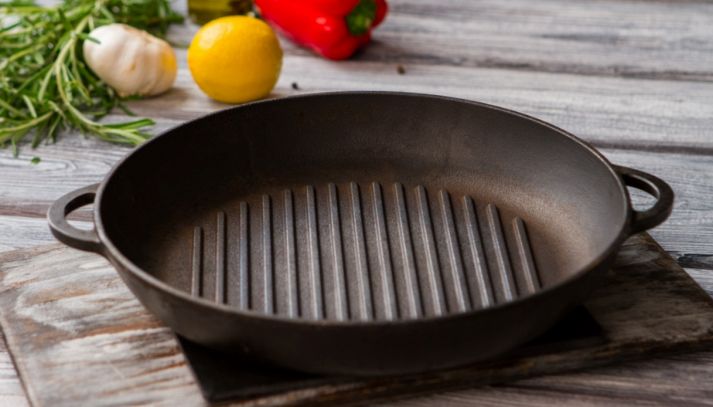 Padella grill rotonda in ghisa con fondo scanalato, posata su un tavolo di legno rustico con rosmarino, aglio, limone e peperone sullo sfondo.