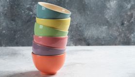 Pila verticale di sei ciotole in ceramica di colori diversi (arancione, viola, rosa, verde, giallo, azzurro) su sfondo grigio testurizzato.