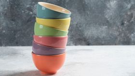 Pila verticale di sei ciotole in ceramica di colori diversi (arancione, viola, rosa, verde, giallo, azzurro) su sfondo grigio testurizzato.