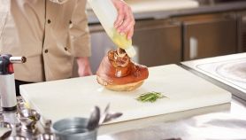 Chef che versa olio su uno stinco arrosto posizionato su un robusto tagliere professionale bianco in polietilene, in una cucina industriale.