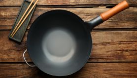 Padella wok per induzione con manico in legno, vuota, posata su un tavolo di legno rustico accanto a un poggia bacchette con due bacchette di legno.