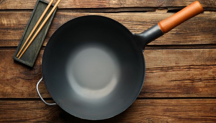 Padella wok per induzione con manico in legno, vuota, posata su un tavolo di legno rustico accanto a un poggia bacchette con due bacchette di legno.