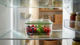 Primo piano di un contenitore trasparente per frigorifero con all'interno frutta e verdura, illuminato da luce naturale.