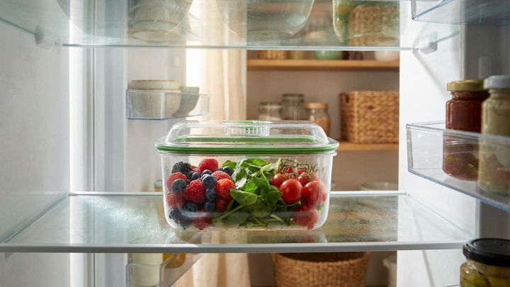 Primo piano di un contenitore trasparente per frigorifero con all'interno frutta e verdura, illuminato da luce naturale.