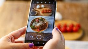 Mani che fotografano un piatto di cibo con uno smartphone su un tavolo, con ingredienti come pomodorini e mozzarella sullo sfondo