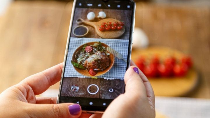 Mani che fotografano un piatto di cibo con uno smartphone su un tavolo, con ingredienti come pomodorini e mozzarella sullo sfondo