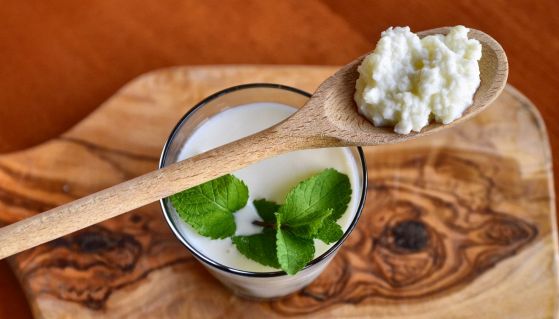 Un cucchiaio di legno solleva dei chicchi di kefir al di sopra di un bicchiere di kefir fresco arricchito con foglie di menta