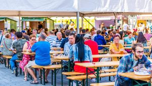 Persone sedute ai tavoli di legno di una sagra all’aperto, che mangiano e bevono sotto un tendone affollato
