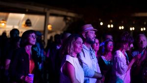 Persone di diverse età assistono a uno spettacolo serale all’aperto, illuminate da luci colorate.