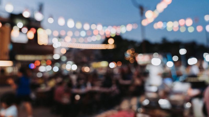 Festa serale all’aperto sfocata, con folla ai tavoli e fili di luci colorate sospesi nel cielo azzurro