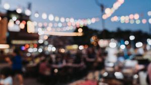 Festa serale all’aperto sfocata, con folla ai tavoli e fili di luci colorate sospesi nel cielo azzurro