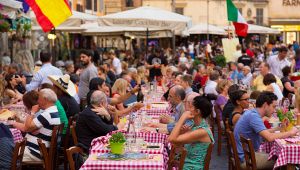 Lunga tavolata all’aperto con tovaglie a quadri, persone che mangiano e chiacchierano tra bandiere e stand della festa