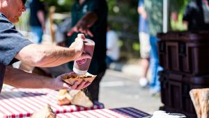 Uomo versa salsa su un vassoio di cibo a un banco con tovaglia a quadri durante una festa all’aperto